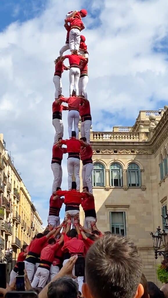 A large group of people in coordinated red shirts and white pants forms a towering human pyramid, known as a "castell," in a city square in Barcelona. The tower rises at least six levels high with a child wearing a red helmet climbing to the top, while a crowd looks on and photographs the event.