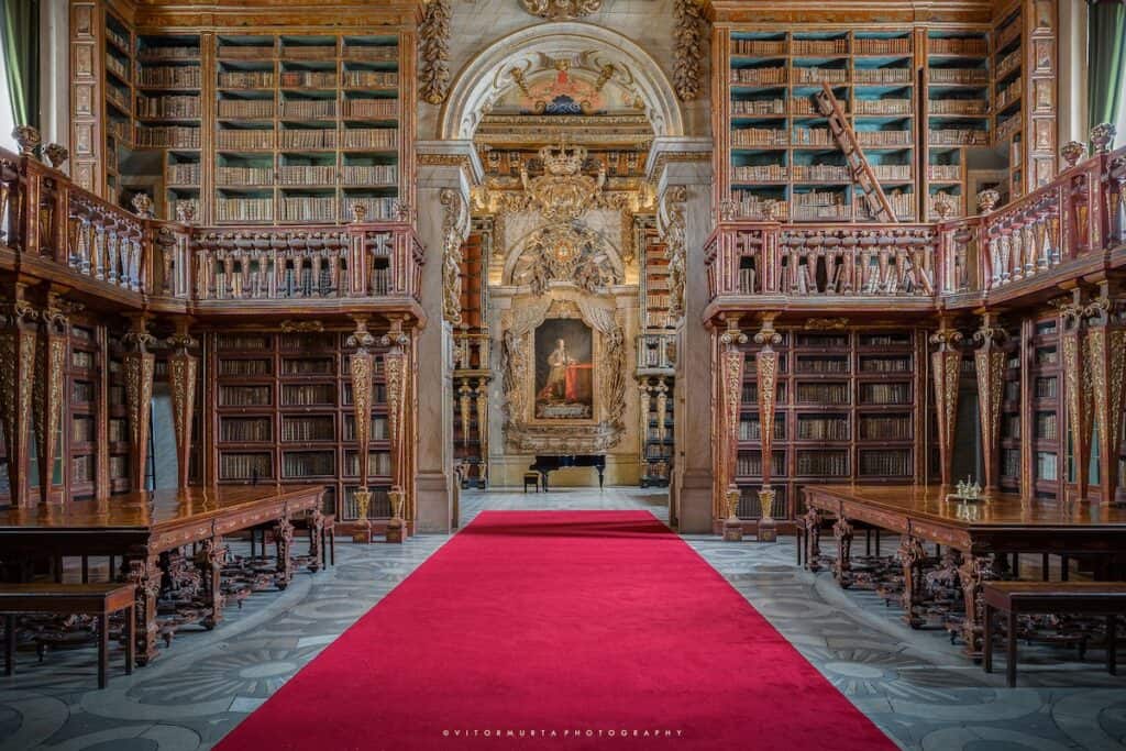 A grand Baroque-style library with towering wooden bookshelves adorned with gold detailing stretches along both sides of a red carpeted aisle. The ornate ceiling and intricate carvings frame a central painting of a regal figure, illuminated beneath an arched alcove. A black grand piano sits at the far end, adding to the elegance of the historic space.