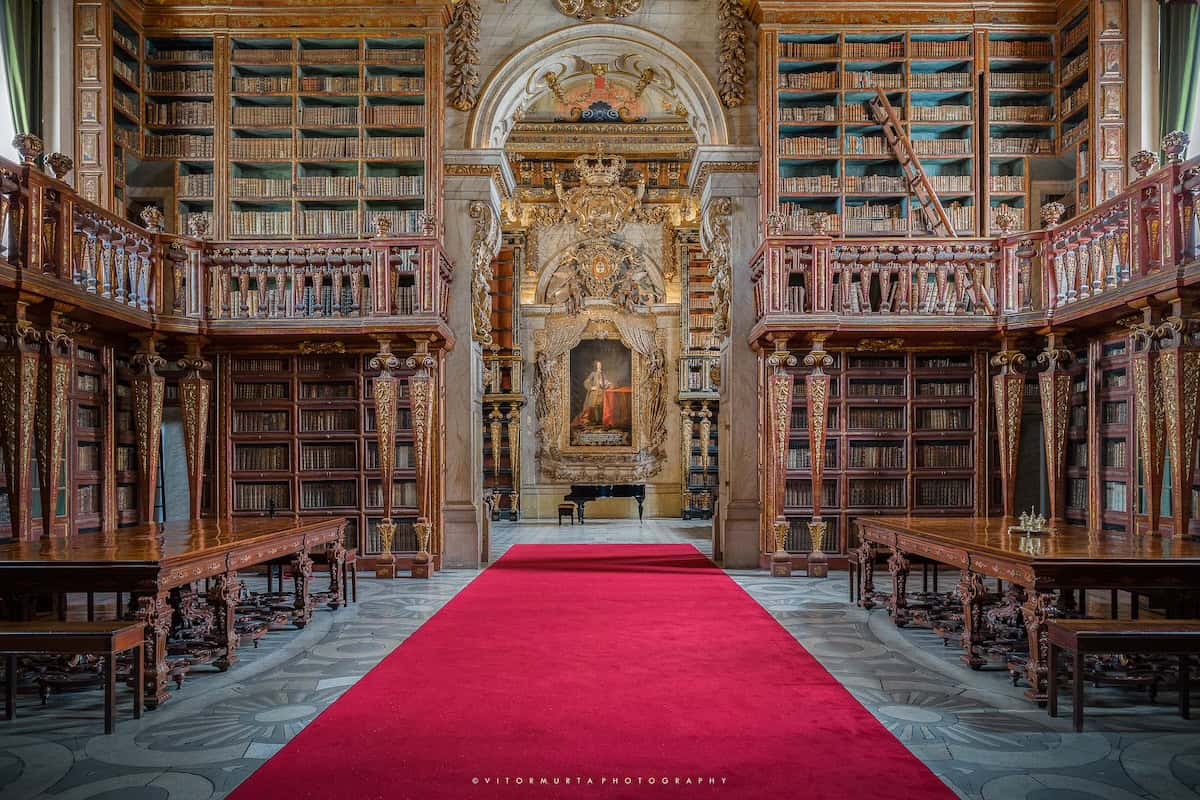 A grand Baroque-style library with towering wooden bookshelves adorned with gold detailing stretches along both sides of a red carpeted aisle. The ornate ceiling and intricate carvings frame a central painting of a regal figure, illuminated beneath an arched alcove. A black grand piano sits at the far end, adding to the elegance of the historic space.