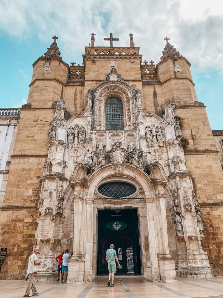 The ornate façade of a historic church, featuring intricate Manueline-style stone carvings, statues, and decorative arches, stands against a partly cloudy sky. The grand entrance, framed by elaborate sculptures, leads into the dimly lit interior. Visitors in casual attire walk towards the entrance, adding a sense of scale to the impressive architecture.