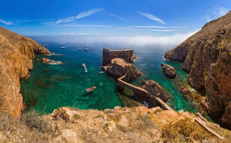 A breathtaking view of the Fortress of São João Baptista on Berlenga Grande, an island off the coast of Portugal. The 17th-century stone fort sits on a rocky outcrop surrounded by crystal-clear turquoise waters, with boats anchored nearby. A narrow stone bridge connects the fortress to the mainland, winding through the rugged, golden cliffs under a bright blue sky.