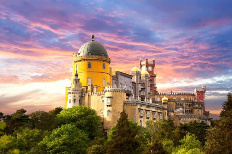 Colorful view of Pena Palace in Sintra, Portugal, with its vibrant yellow, red, and lavender towers rising above lush green trees at sunset. The sky is painted with dramatic hues of pink, purple, and gold, adding a magical backdrop to the Romanticist castle's whimsical architecture.