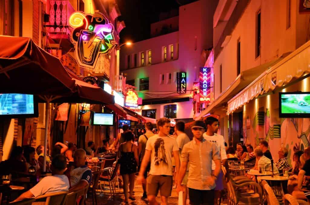 Vibrant nightlife scene in Albufeira, Portugal, showing a bustling street lined with neon-lit bars, karaoke signs, and outdoor cafés packed with people. Groups of tourists stroll past tables as others watch sports on outdoor TVs under warm lighting and colorful signage.