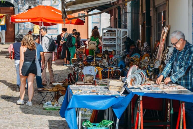 A lively scene at Feira da Ladra, the famous flea market in Lisbon, Portugal. Various antique and vintage items, including ceramic plates, postcards, old books, and decorative trinkets, are displayed on tables covered with blue cloths. A man in a plaid shirt examines some postcards, while shoppers browse the stalls and walk along the cobbled street under the shade of red umbrellas.