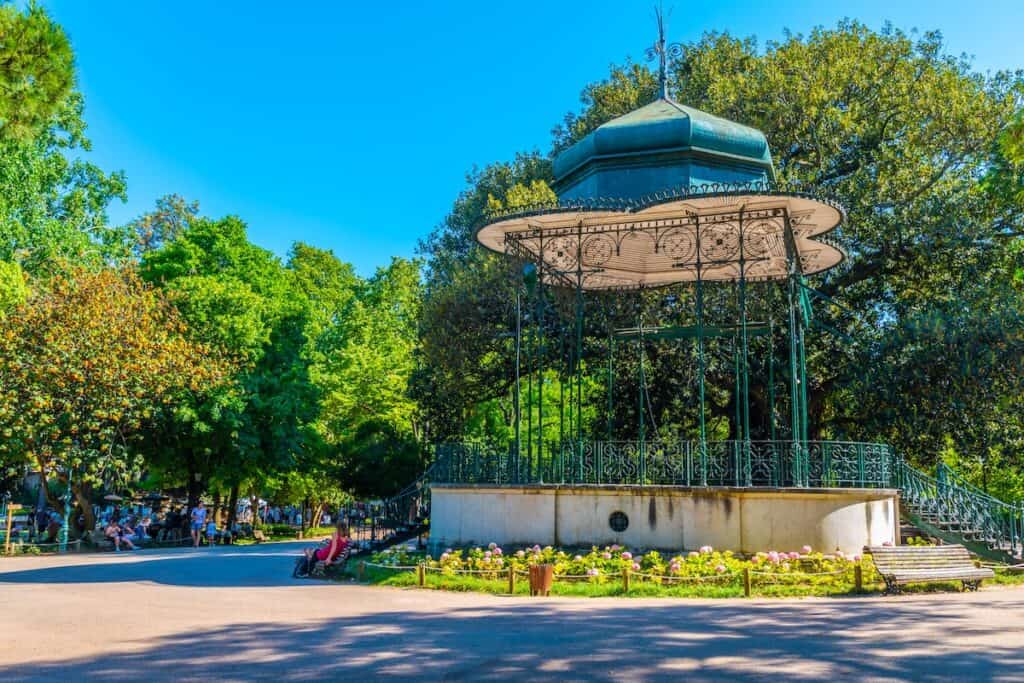 A beautiful iron gazebo with intricate detailing stands in Jardim da Estrela, a lush public park in Lisbon, Portugal. The gazebo is surrounded by vibrant flower beds and towering trees, creating a peaceful and inviting atmosphere. Sunlight filters through the greenery as people relax on benches and stroll along the pathways in the background.