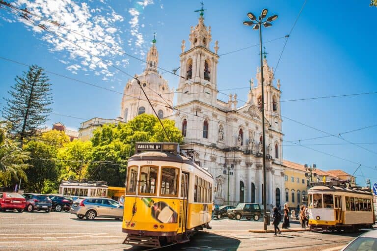 Bright yellow vintage trams in Lisbon, Portugal, with the white Basílica da Estrela in the background under a clear blue sky. The scene includes pedestrians, parked cars, and overhead tram wires crisscrossing the sunny street.