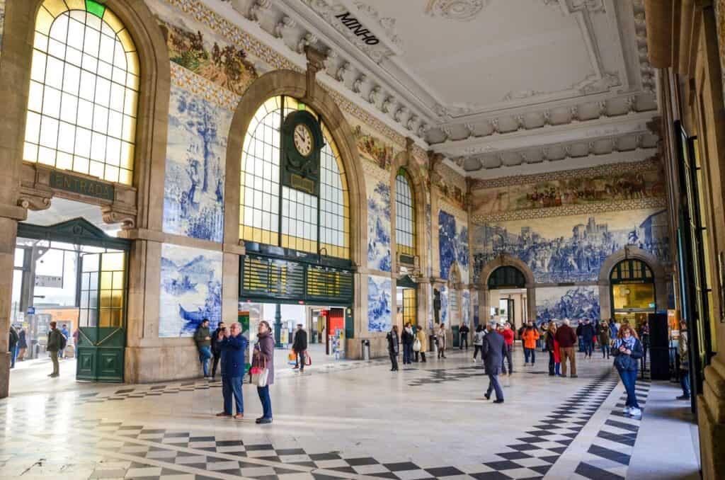 Interior of São Bento Railway Station in Porto, Portugal, featuring large arched windows, a central clock, and iconic blue-and-white azulejo tile murals depicting historical scenes. Travelers and tourists are scattered around the ornate hall, admiring the artwork and checking schedules.