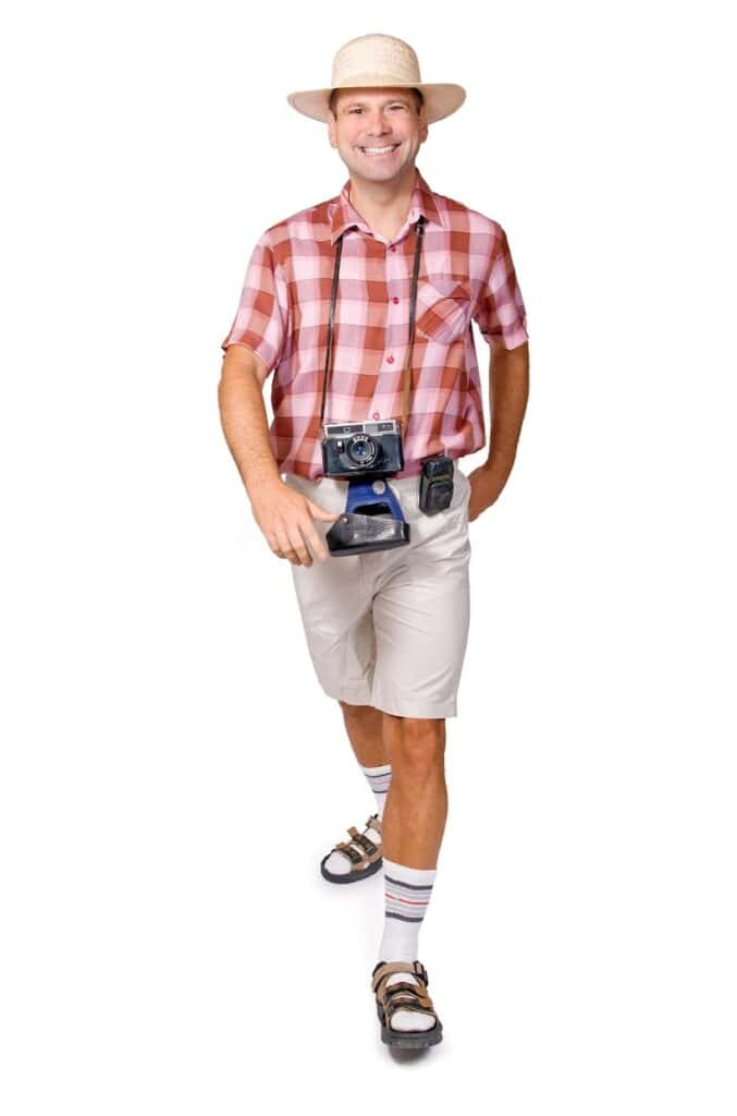 Smiling man dressed like a stereotypical tourist, wearing a straw hat, red plaid shirt, beige shorts, and socks with sandals. He has an old camera around his neck and a fanny pack, striking a cheerful pose on a white background.