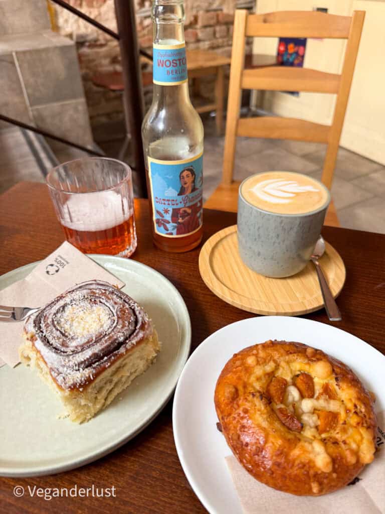 A cozy café table spread featuring a cinnamon roll, an almond Danish pastry, a foamy latte with latte art, and a bottle of Wostok pomegranate-date soda. The rustic setting adds to the laid-back, homey vibe.