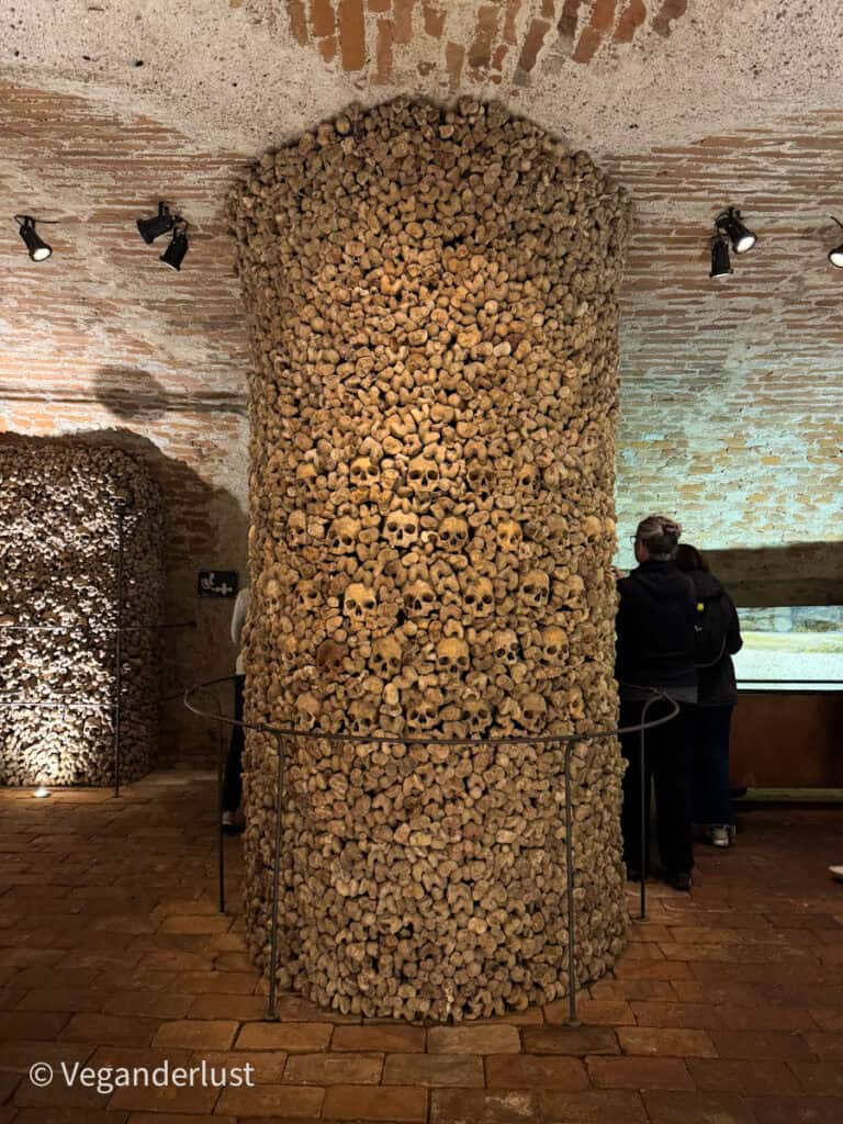 A cylindrical pillar entirely constructed from human skulls and bones stands in a dimly lit brick-vaulted chamber of the Brno Ossuary. Visitors observe the macabre yet structured arrangement, which reflects historical practices of honoring the dead in a confined sacred space.