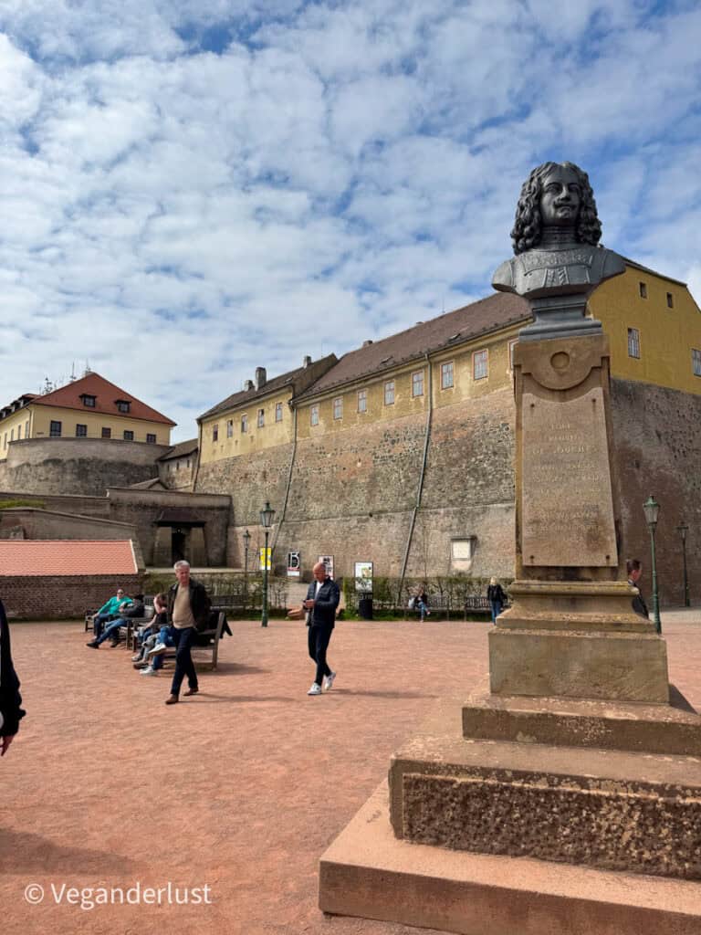 A bronze bust of Louis Raduit de Souches is displayed on a pedestal in the courtyard of Špilberk Castle. Visitors walk around the reddish gravel square with the fortress walls and yellow buildings in the background.