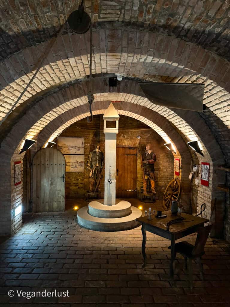 An underground chamber set up as a medieval torture or justice display, featuring a central pillar with restraints, mannequins in historical dress, and a noose hanging from the ceiling.