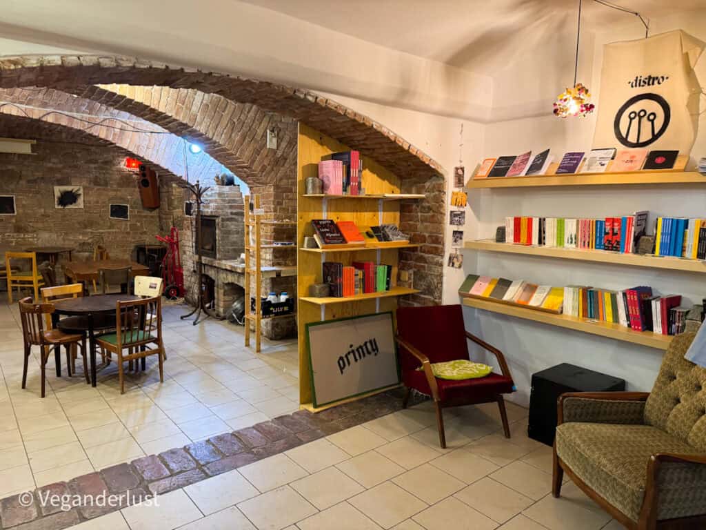 Cozy interior of a vegan-friendly café and community space with exposed brick arches, mismatched wooden tables and chairs, and colorful books arranged on wooden shelves; armchairs and soft lighting add a homey, relaxed vibe.