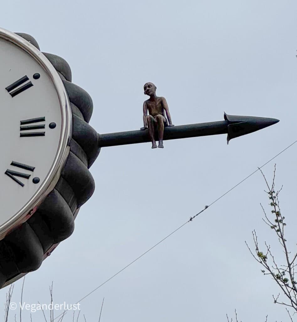 A whimsical sculpture of a small seated figure perches on the end of a clockโs minute hand, high above the street. The childlike statue adds a playful twist to the cityโs skyline.