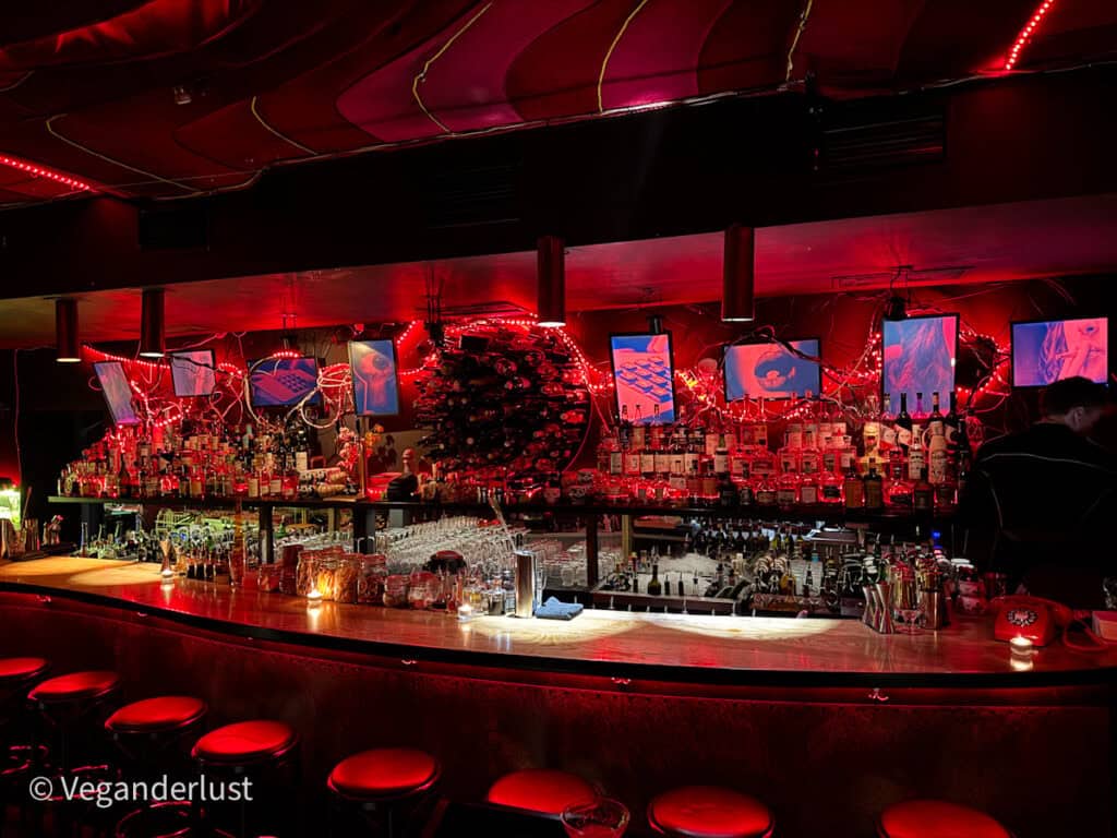 A moody, neon-lit cocktail bar with deep red lighting and an eclectic decor. The long wooden bar is lined with red stools and bottles, while multiple TV screens and string lights create a surreal, circus-inspired vibe—this is the iconic Super Panda Circus in Brno.
