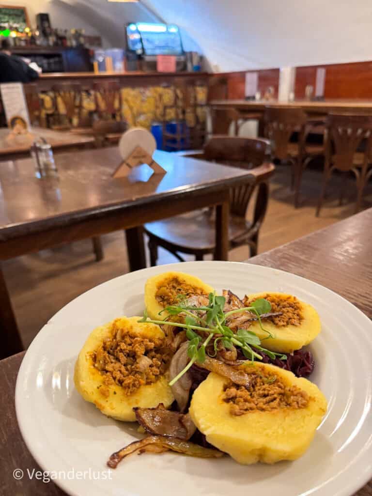 A traditional Czech dish gets a vegan twist—five fluffy potato dumplings filled with savory lentil mixture, served with caramelized onions, fresh herbs, and red cabbage on a white plate inside a casual pub setting.