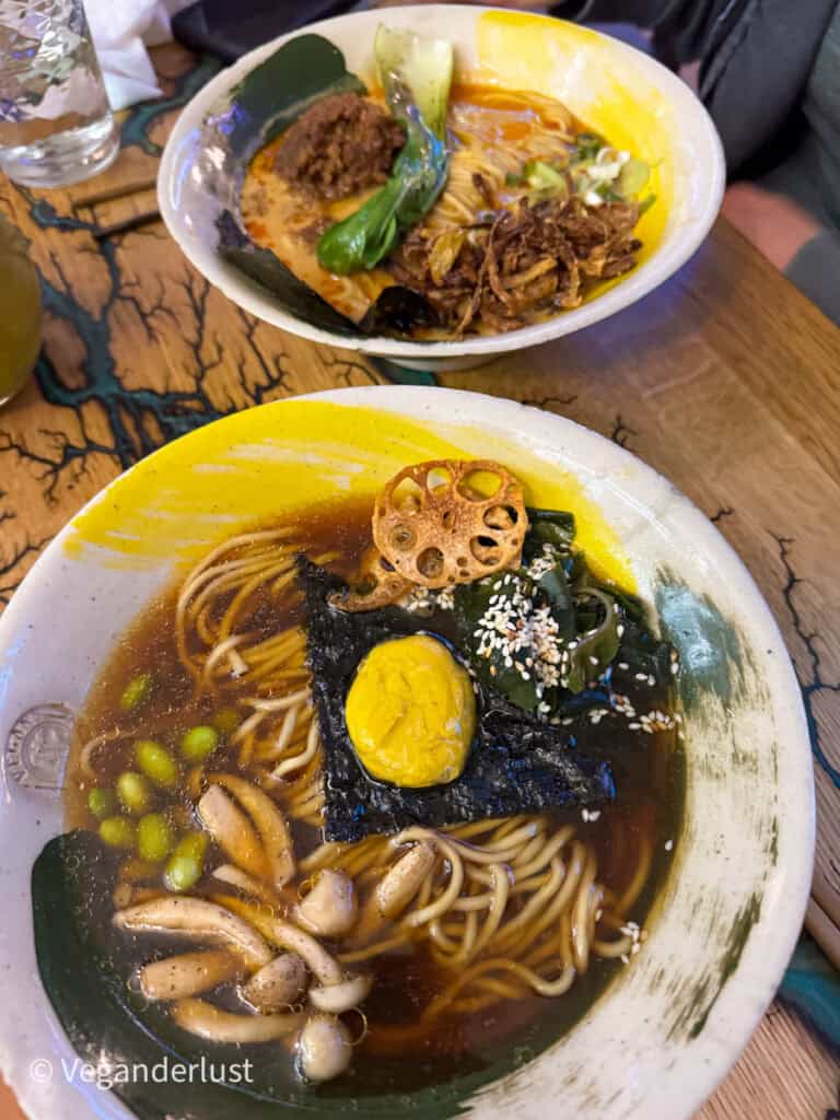Two colorful bowls of vegan ramen packed with toppings—shiitake mushrooms, seaweed, edamame, lotus root, bok choy, and a bright yellow egg substitute—set on a vibrant, rustic wooden table.