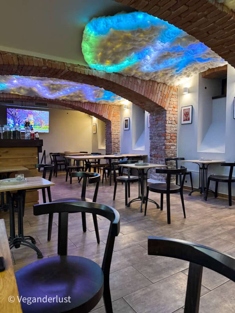 Interior of a modern ramen restaurant with exposed brick arches and whimsical cloud-like ceiling lights glowing in shades of blue, purple, and yellow; simple wooden tables and black chairs are arranged neatly across the tiled floor.