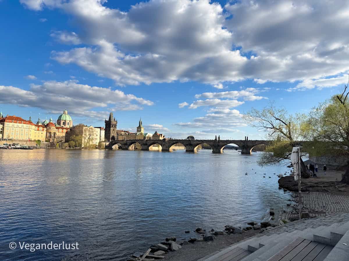 View of the iconic Charles Bridge in Prague spanning the Vltava River, with historic buildings and church domes on the left under a sky filled with fluffy clouds.