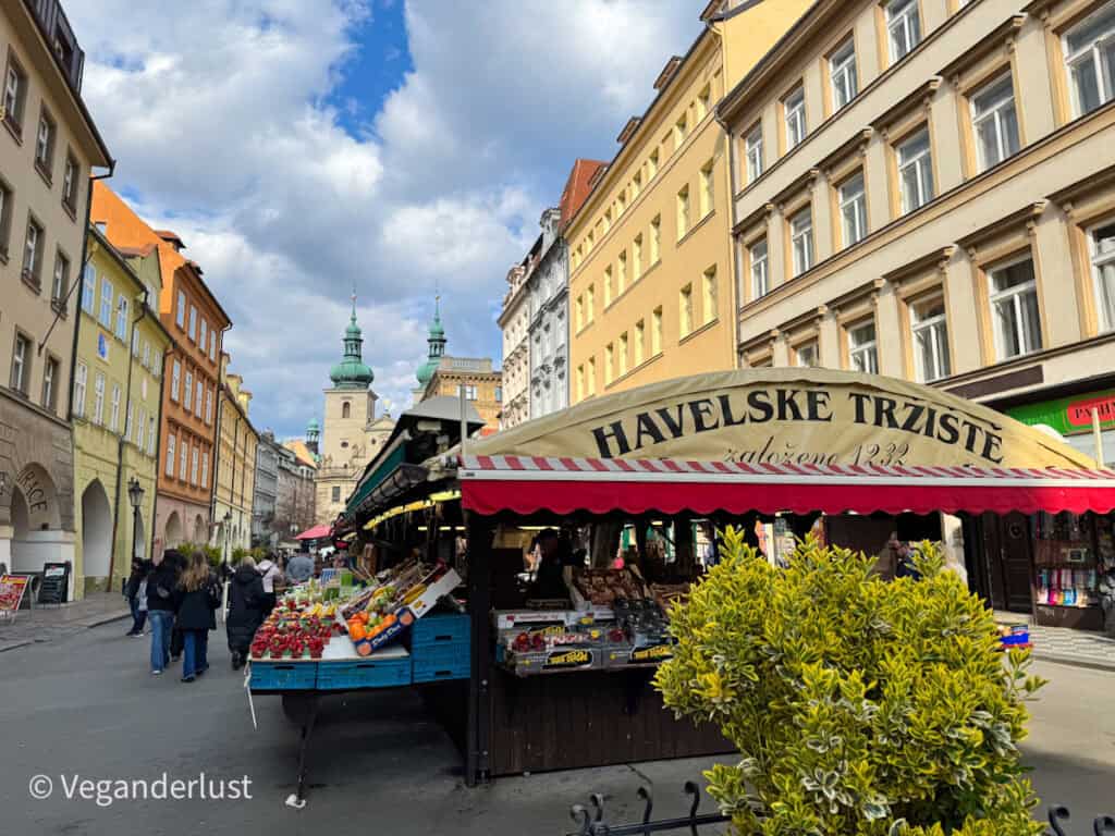 A vibrant street view of Havel’s Market (Havelské Tržiště) in Prague, with colorful produce and souvenirs displayed under a red-and-white striped canopy. Historic buildings and church towers line the bustling pedestrian street under a partly cloudy sky.