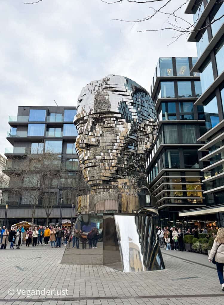 A crowd gathers around the massive kinetic sculpture of Franz Kafka’s rotating head, made of shiny mirrored layers, surrounded by modern glass buildings in Prague.
