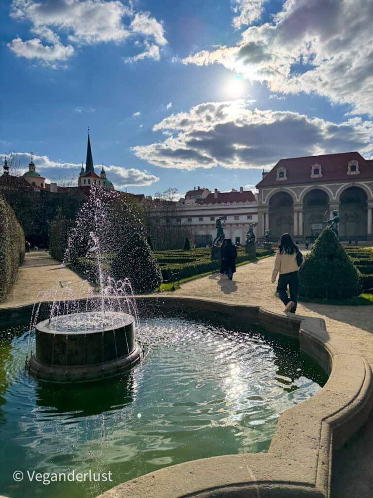 A peaceful view of Waldstein Garden in Prague, with a stone fountain spraying water in the foreground, neatly trimmed hedges, and baroque sculptures lining the paths. The sun shines through scattered clouds, casting a golden light over the scene.