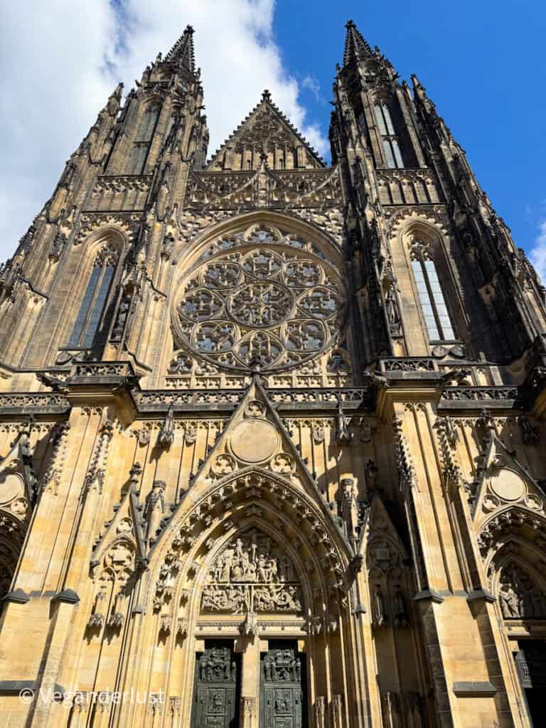 The grand Gothic facade of St. Vitus Cathedral in Prague, towering with ornate spires and a large rose window. The stonework is intricately detailed, glowing warmly under the sunlight and blue sky.