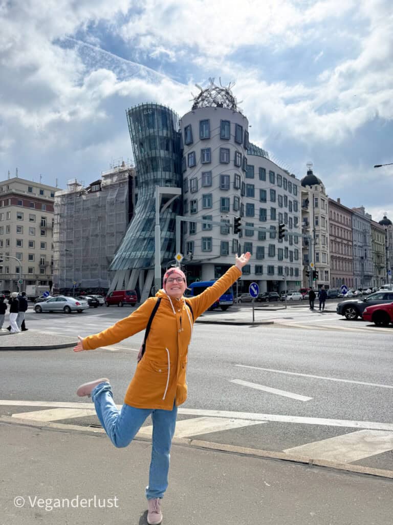 Tina in a mustard yellow jacket strikes a playful pose in front of Prague’s famous Dancing House, a modern architectural landmark with curvy lines and contrasting glass and concrete towers. The sky is partly cloudy, and the street around her is lively with cars and pedestrians.