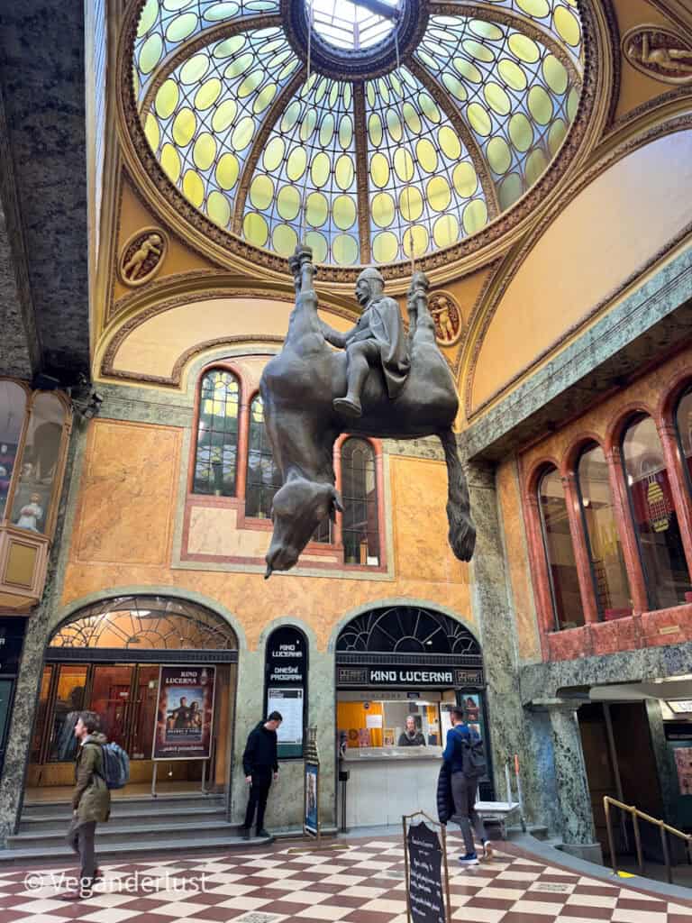 Inside Prague’s Lucerna Palace, a surreal sculpture of Saint Wenceslas riding an upside-down dead horse hangs from the ceiling beneath an ornate stained-glass dome. The bizarre statue contrasts with the art nouveau elegance of the surrounding cinema lobby.
