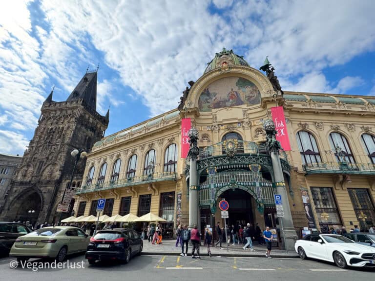 The ornate facade of Prague’s Municipal House with its decorative archway, mosaic, and sculptures, next to the dark gothic Powder Tower. People and cars fill the busy square out front.