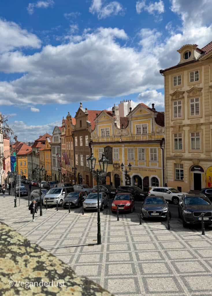 A picturesque cobblestone street in Prague lined with pastel-colored historic buildings, red-tiled roofs, and vintage street lamps under a vibrant blue sky with dramatic clouds. Parked cars and a few pedestrians add a lively touch to this charming European scene.