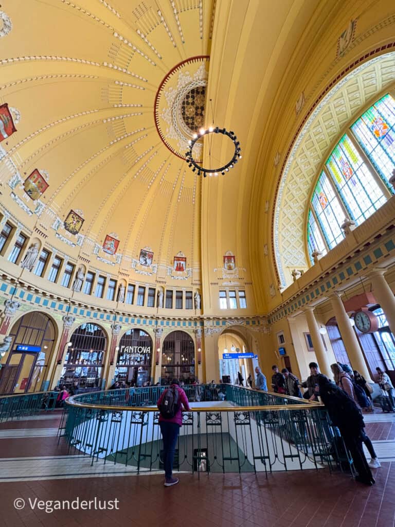 Inside the art nouveau dome of Prague’s main train station, Fantova kavárna is visible with people gathered under the grand yellow ceiling adorned with coats of arms and intricate moldings.