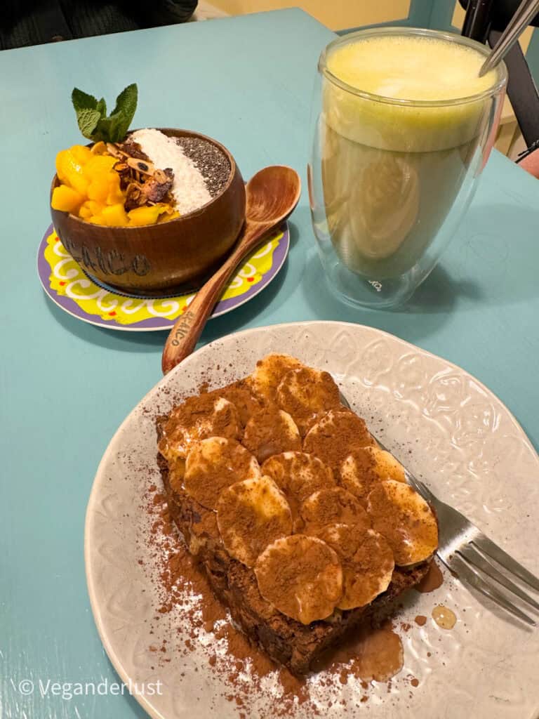 A vibrant vegan breakfast spread featuring a smoothie bowl with mango, chia seeds, coconut, and granola in a wooden bowl, paired with banana chocolate toast dusted with cocoa powder and a tall green smoothie on a turquoise table.