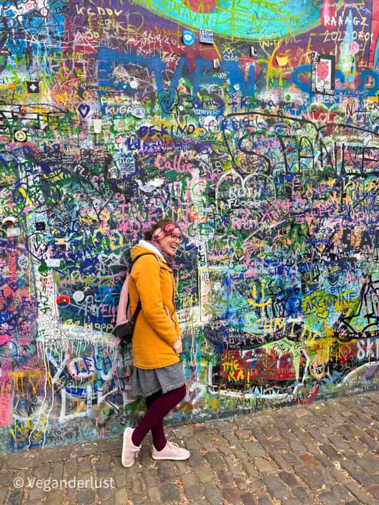 Tina in a mustard yellow coat and pink headband stands in front of Prague’s vibrant Lennon Wall, covered in colorful graffiti, peace signs, and messages like “Be Kind” and “LOVE.” The cobblestone path beneath her adds to the artsy, bohemian vibe.
