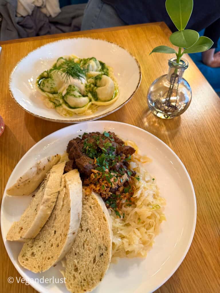 A plate of traditional Czech vegan dumplings served with cabbage and a plant-based meat stew, next to another bowl of dumplings topped with green herb sauce, at a rustic wooden table.