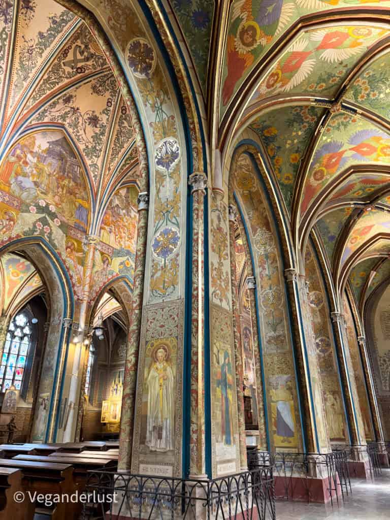 Inside the richly decorated Vyšehrad church in Prague, showcasing arched columns and vaulted ceilings covered in intricate, colorful murals of saints, angels, and floral patterns. Sunlight filters through tall stained glass windows, adding to the serene atmosphere.