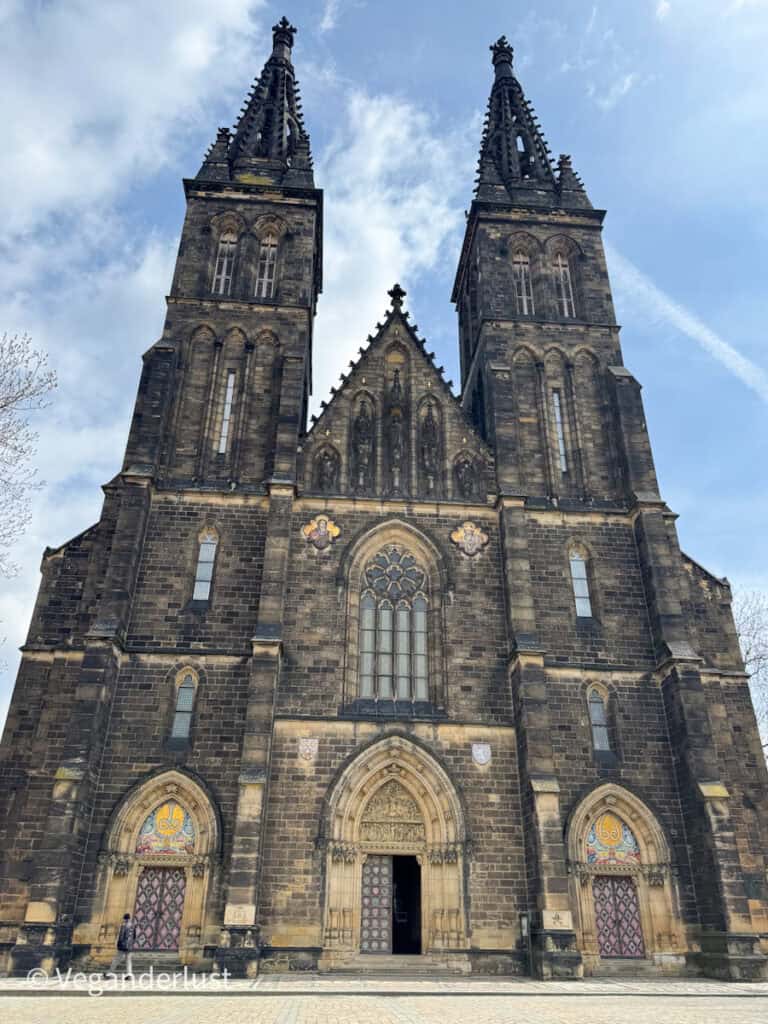 The front view of the Basilica of St. Peter and St. Paul in Vyšehrad, Prague, showcasing its dark Gothic twin towers and intricately decorated entry doors. The sky above is partly cloudy, adding contrast to the dramatic stone facade.