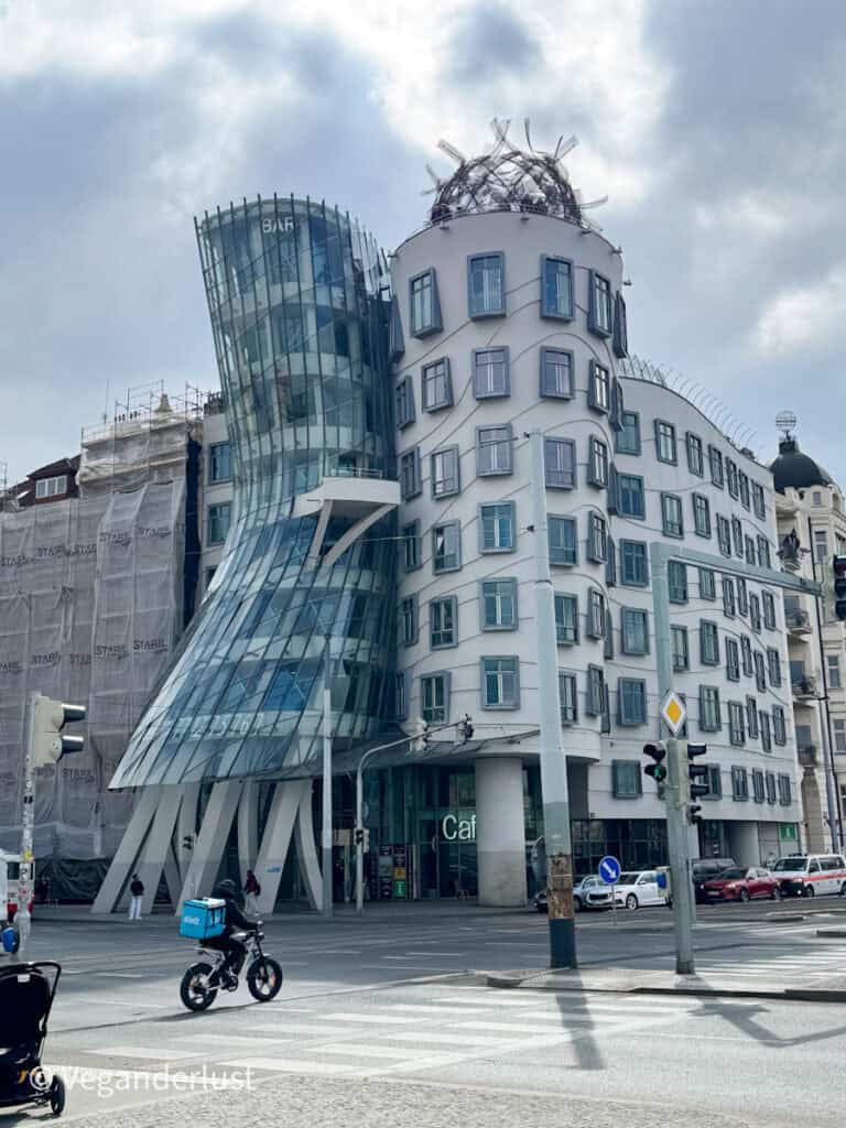 The quirky, modern architecture of the Dancing House in Prague, with its curved glass and concrete towers, stands out at a street intersection. A food delivery cyclist rides past the building, adding a sense of city life.