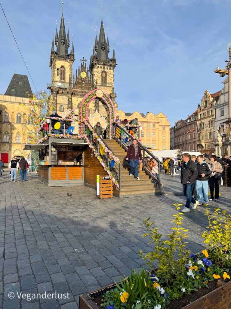 A festive Easter market setup in Prague’s Old Town Square features a wooden platform with colorful flower garlands and decorative eggs leading up to a viewing deck. The Church of Our Lady before Týn stands tall in the background under a clear blue sky.