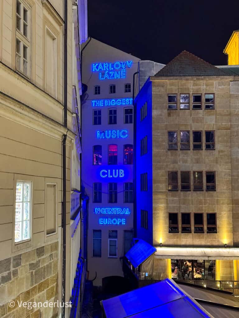 Nighttime view of Karlovy Lázně, a multi-story building in Prague lit up with bright blue neon signs reading "The biggest music club in Central Europe." The narrow alleyway and glowing windows below hint at the city's vibrant nightlife scene.