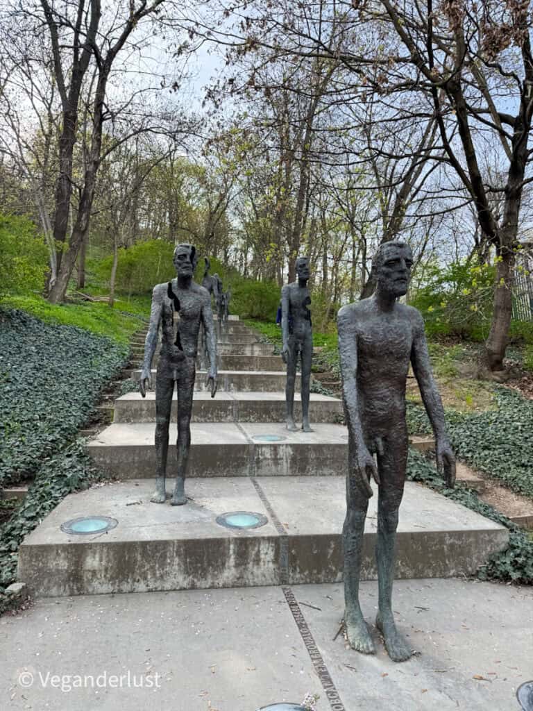 A series of haunting bronze male figures descend a flight of stairs in a wooded park, each progressively more fragmented, symbolizing the physical and psychological toll of totalitarian rule in the Czech Republic.