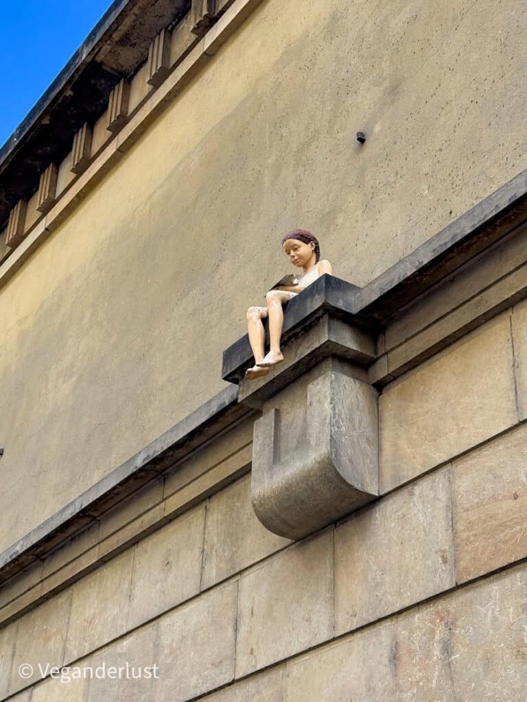 A lifelike sculpture of a young girl sits barefoot on a stone ledge high up on a beige building wall, reading a book with a peaceful expression under a clear blue sky.