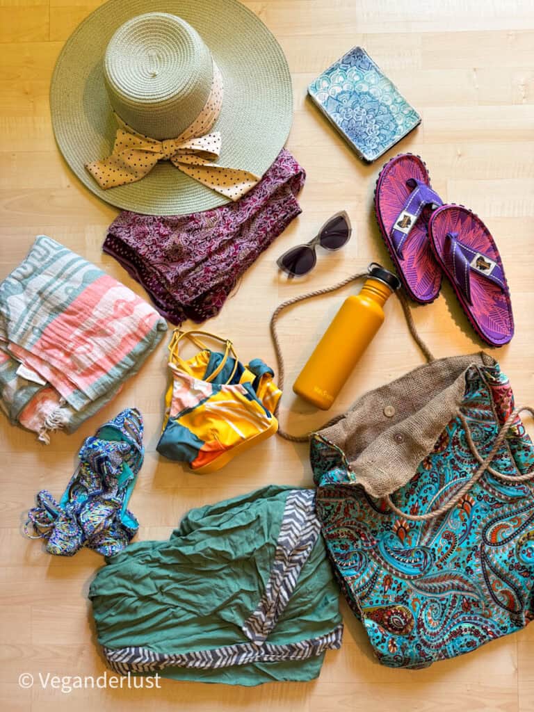 Flat lay of summer beach essentials on a wooden floor, including a wide-brim straw hat with a polka-dot ribbon, a folded sarong, sunglasses, a yellow water bottle, sandals, two bikinis, a striped towel, and a large paisley print beach bag.