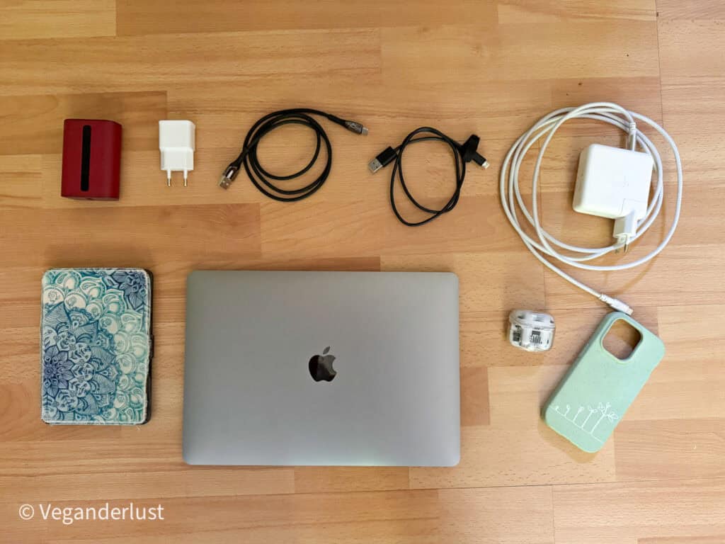 Travel electronics flat lay including a silver MacBook, phone charger, two USB cables, wall adapters, a red power bank, a phone with a mint-green case, and a blue-and-white patterned travel pouch.