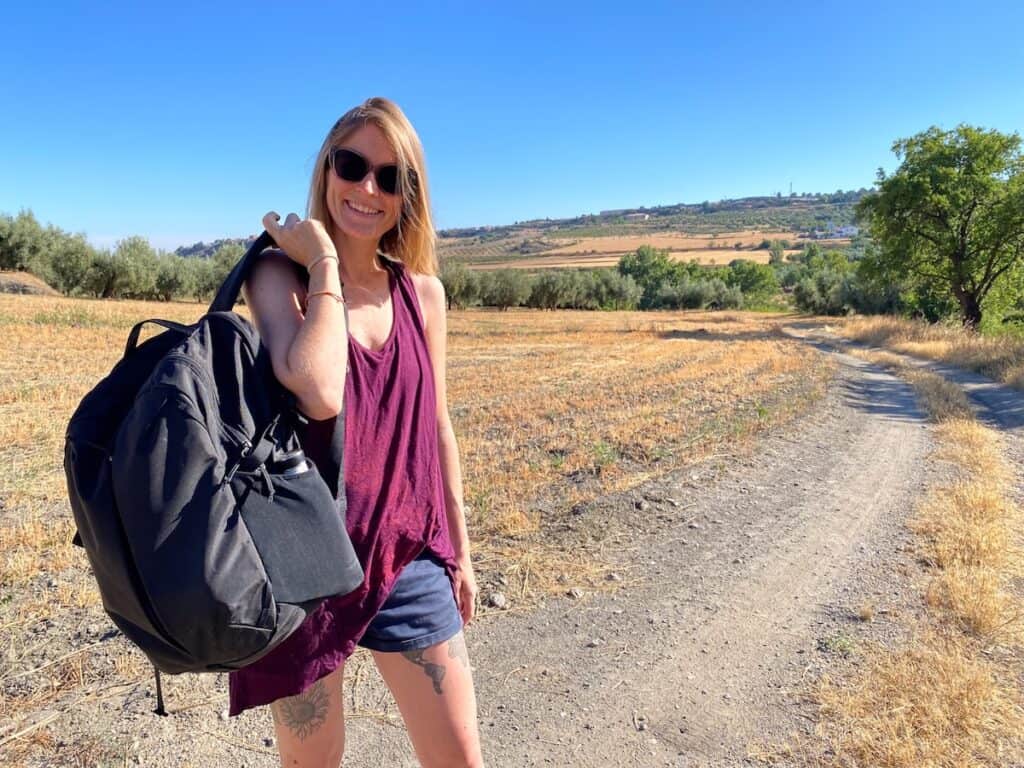 Tina in a sleeveless burgundy top and denim shorts stands on a sunlit dirt trail through a dry, rural landscape. She carries a large black backpack over one shoulder and wears dark sunglasses, with open fields and distant hills behind her.