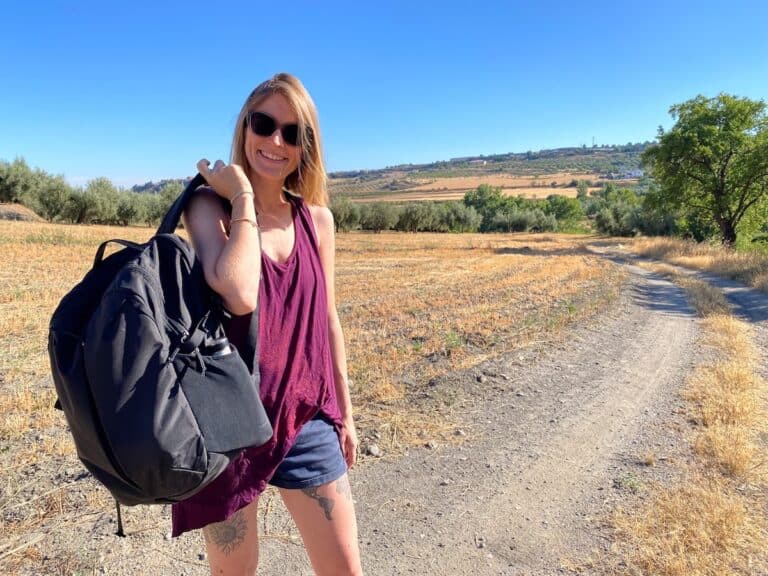 Tina in a sleeveless burgundy top and denim shorts stands on a sunlit dirt trail through a dry, rural landscape. She carries a large black backpack over one shoulder and wears dark sunglasses, with open fields and distant hills behind her.