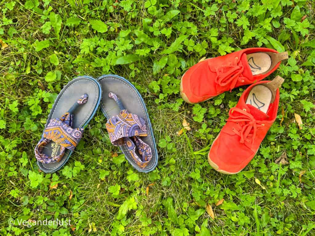 Two pairs of shoes on green grass: colorful patterned sandals with toe loops on the left and bright red minimalist sneakers on the right.
