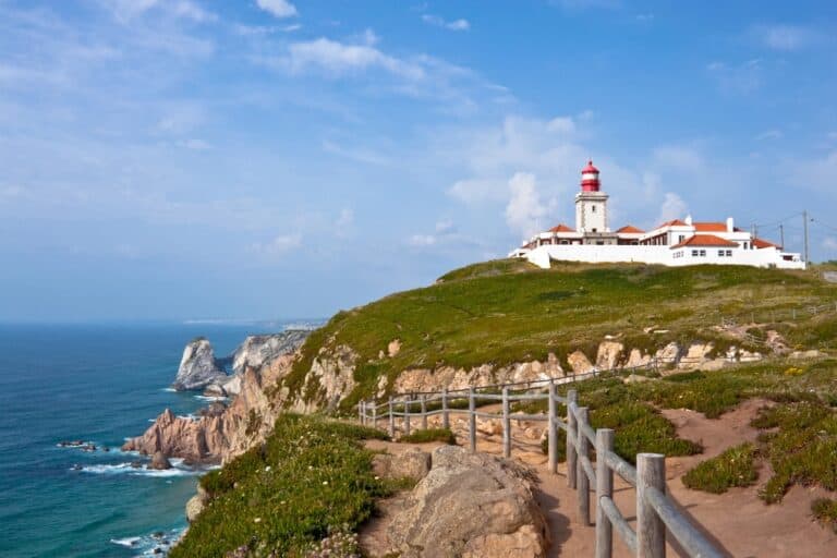 Scenic view of Cabo da Roca in Portugal, featuring a red-roofed lighthouse perched on a grassy cliff overlooking the Atlantic Ocean. A wooden path winds along the rugged coastline, with dramatic sea cliffs and crashing waves below.
