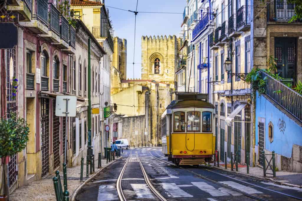Vintage yellow tram traveling through a narrow, cobbled street in Lisbon’s Alfama district, flanked by pastel buildings and leading toward the historic Lisbon Cathedral in the background.