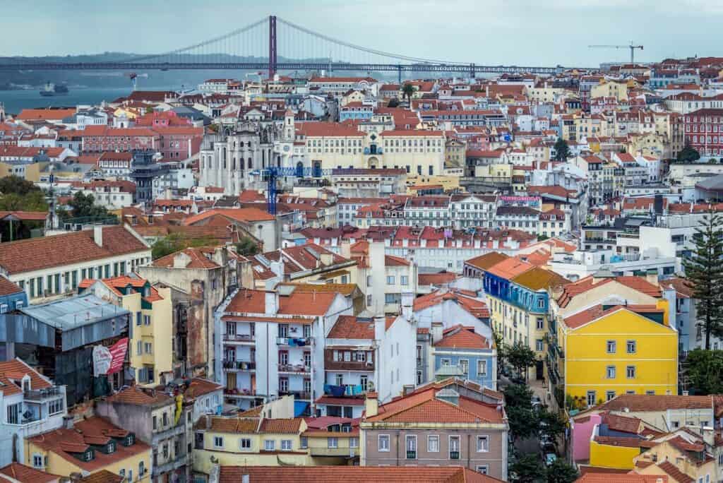 Panoramic view of Lisbon’s historic center with a sea of red-tiled rooftops, colorful buildings, and the 25 de Abril Bridge in the background spanning the Tagus River.